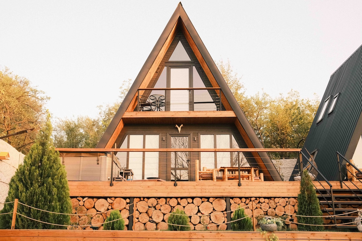 The A-frame cabin features a striking triangular roof and a prominent wooden deck. Stacked logs are positioned below the balcony, providing a rustic charm. Large windows offer views of the surrounding greenery, while outdoor seating is arranged on the terrace.