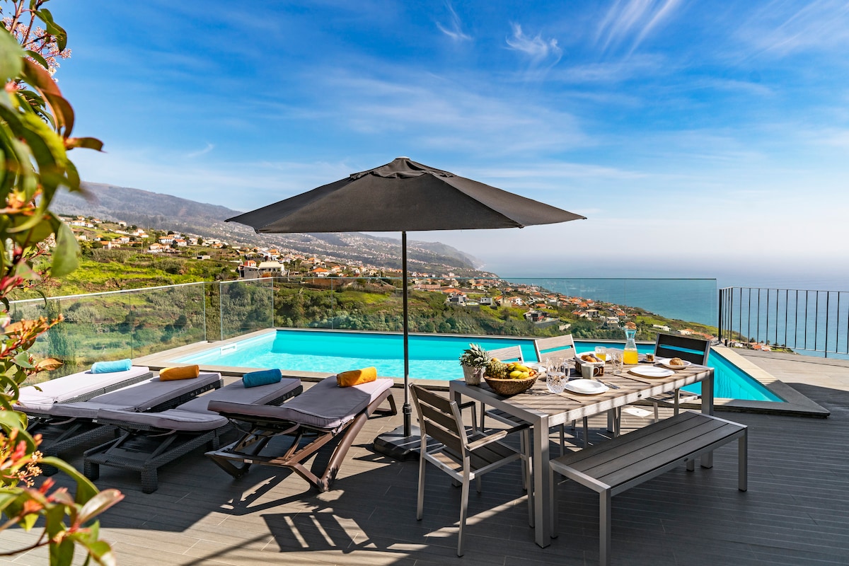 An outdoor terrace features a dining table set for meals and several sun loungers positioned beside a heated infinity pool. Overhead, a large umbrella provides shade, while panoramic views of the Atlantic Ocean and surrounding hills create a scenic backdrop.