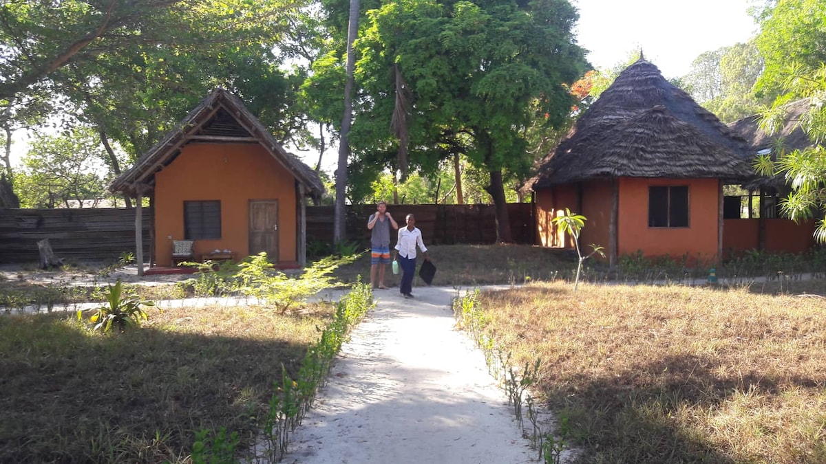 A sandy pathway leads through a lush garden towards two distinct thatched-roof cottages. One cottage features warm orange walls and a window, while the other showcases a simple design. Green foliage surrounds the area, contributing to a serene atmosphere.