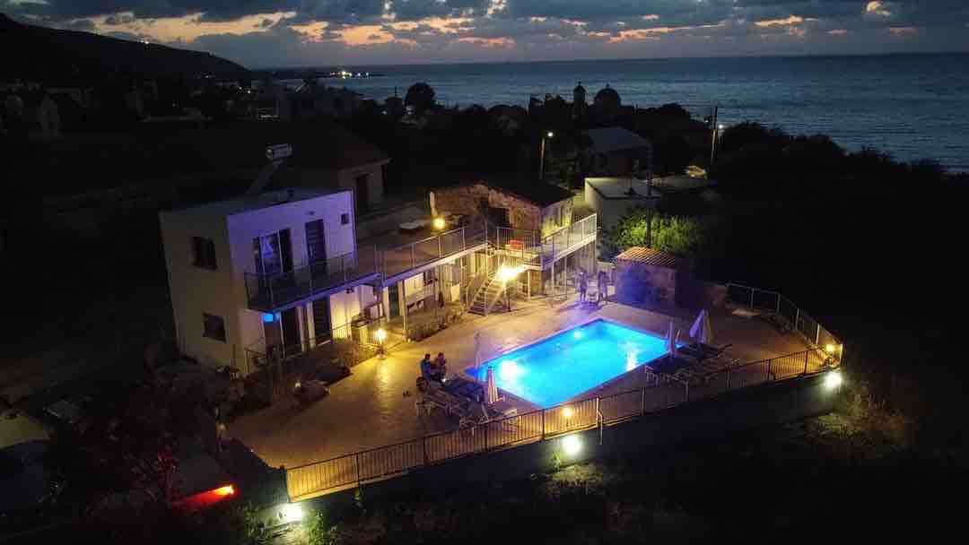 An aerial view of a two-story property showcases an illuminated swimming pool surrounded by lounge chairs. Soft lighting highlights the outdoor space, while the distant coastline and hills are visible against the evening sky. The area is enclosed with a fence for privacy.