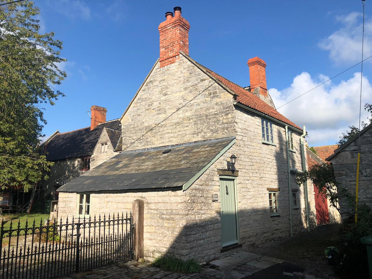 The cottage features a traditional stone exterior with a gabled roof, complemented by distinctive red brick chimneys. A wooden door with a light blue finish is situated on the front, while a small garden area and picket fence enhance the property's charm.