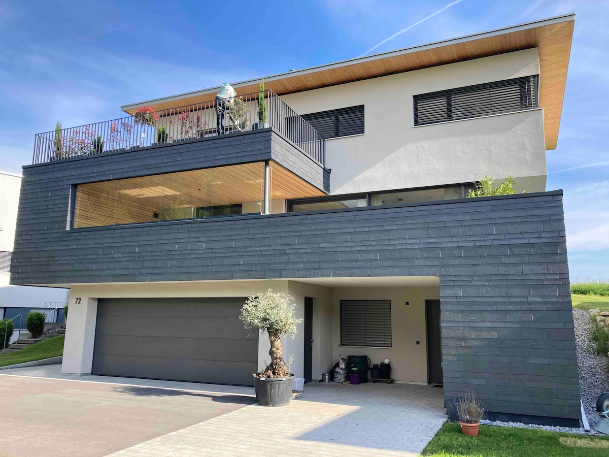 A modern two-story house is presented, featuring a striking combination of dark exterior panels and wooden accents. Balconies with greenery overlook the entrance, while a spacious garage is visible below. A well-maintained driveway leads to the home's entrance.