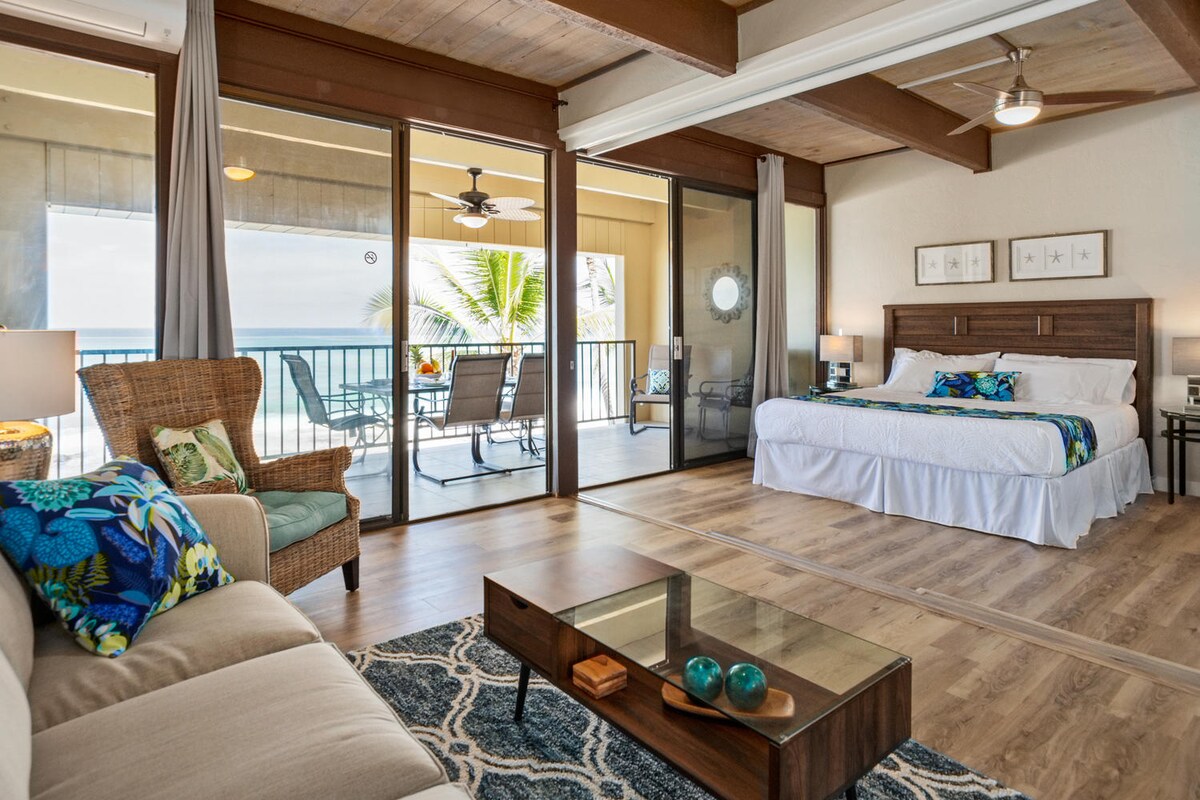 An open bedroom area features a spacious bed adorned with colorful bedding. Natural light filters through large windows, revealing an inviting lanai with seating. A comfortable sofa and a coffee table apply warmth to the space, complemented by a decorative area rug.