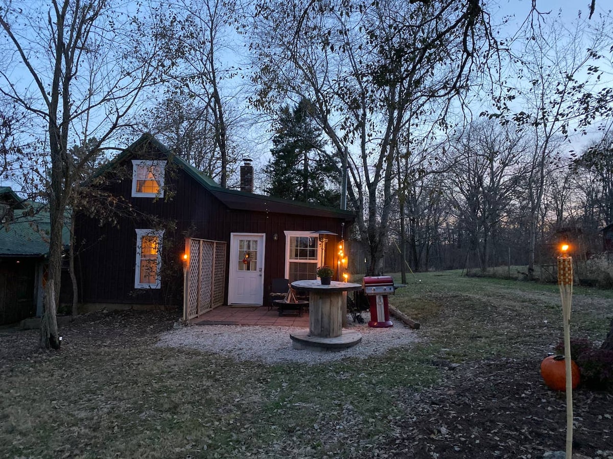 A charming cottage is viewed from the side, featuring a private patio with outdoor seating and a grill. Soft lights illuminate the exterior, surrounded by trees and a grassy area, creating a cozy atmosphere as dusk approaches.