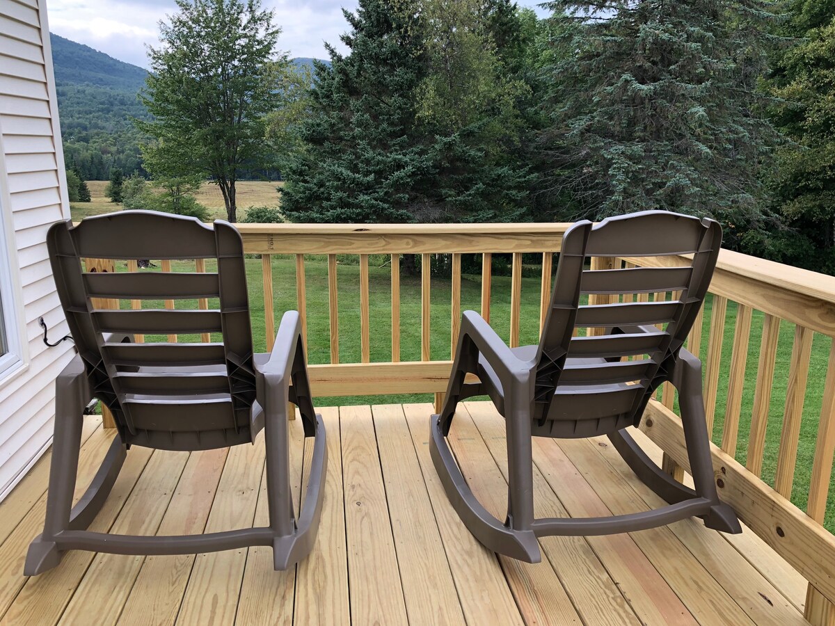 Two rocking chairs are positioned on a wooden deck, facing a scenic view of rolling hills and trees. The deck features a simple railing, complementing the natural surroundings and offering a peaceful spot for outdoor relaxation.