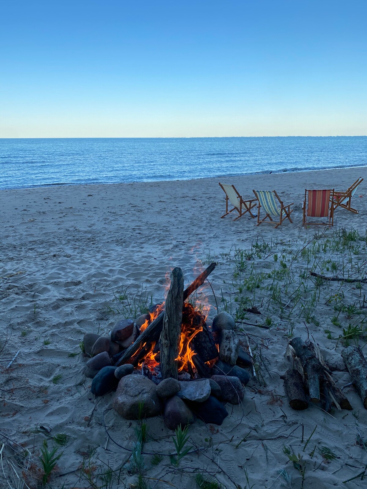 A firepit is surrounded by smooth stones, with flames gently rising in the center. In the background, calm waters of Lake Huron stretch toward the horizon, while a sandy beach leads to a few wooden chairs positioned for relaxation by the shore.