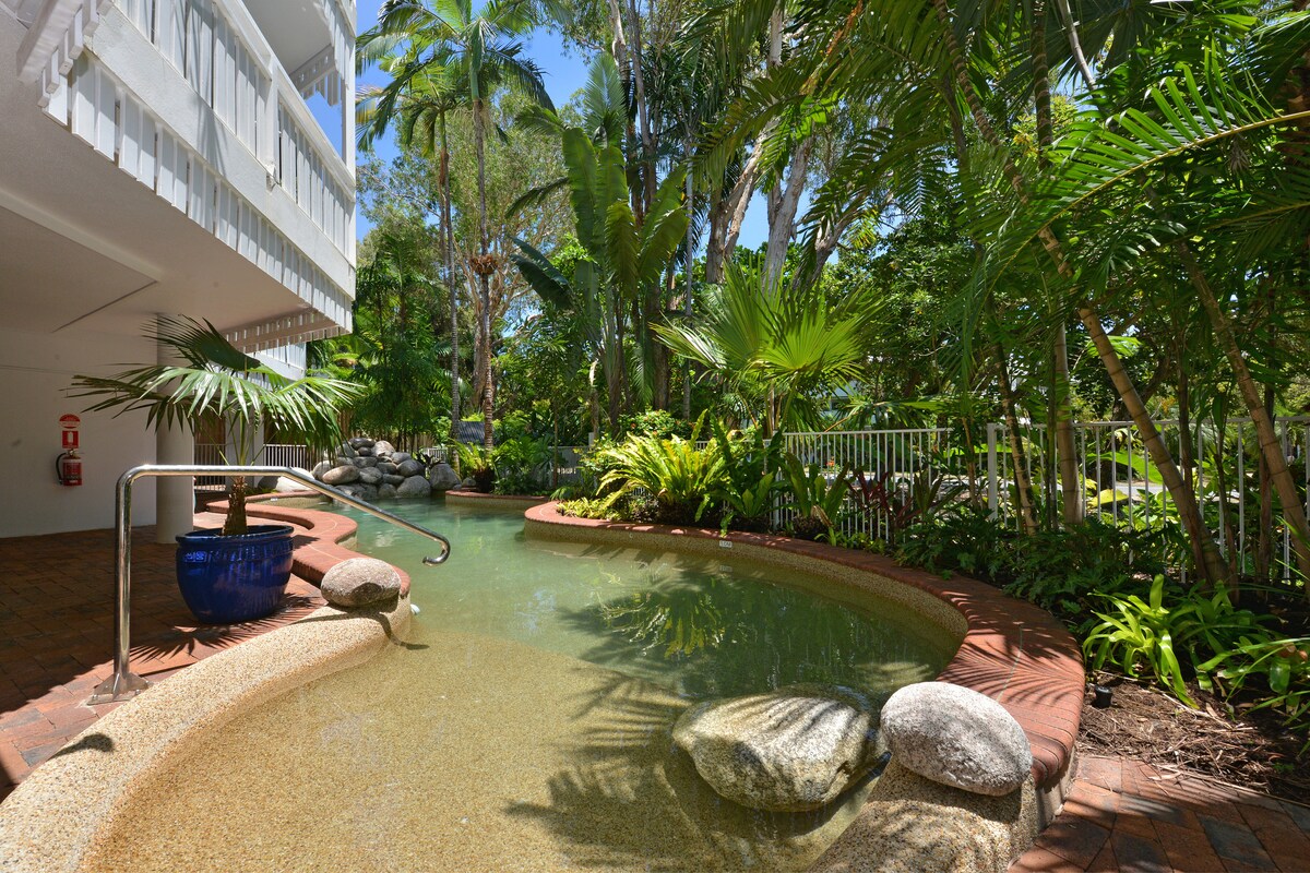 A tranquil shared heated pool is surrounded by lush tropical gardens, featuring various plants and palm trees. The pool area is accented by natural stones and a curved edge, with steps leading into the water, providing a serene atmosphere for relaxation.