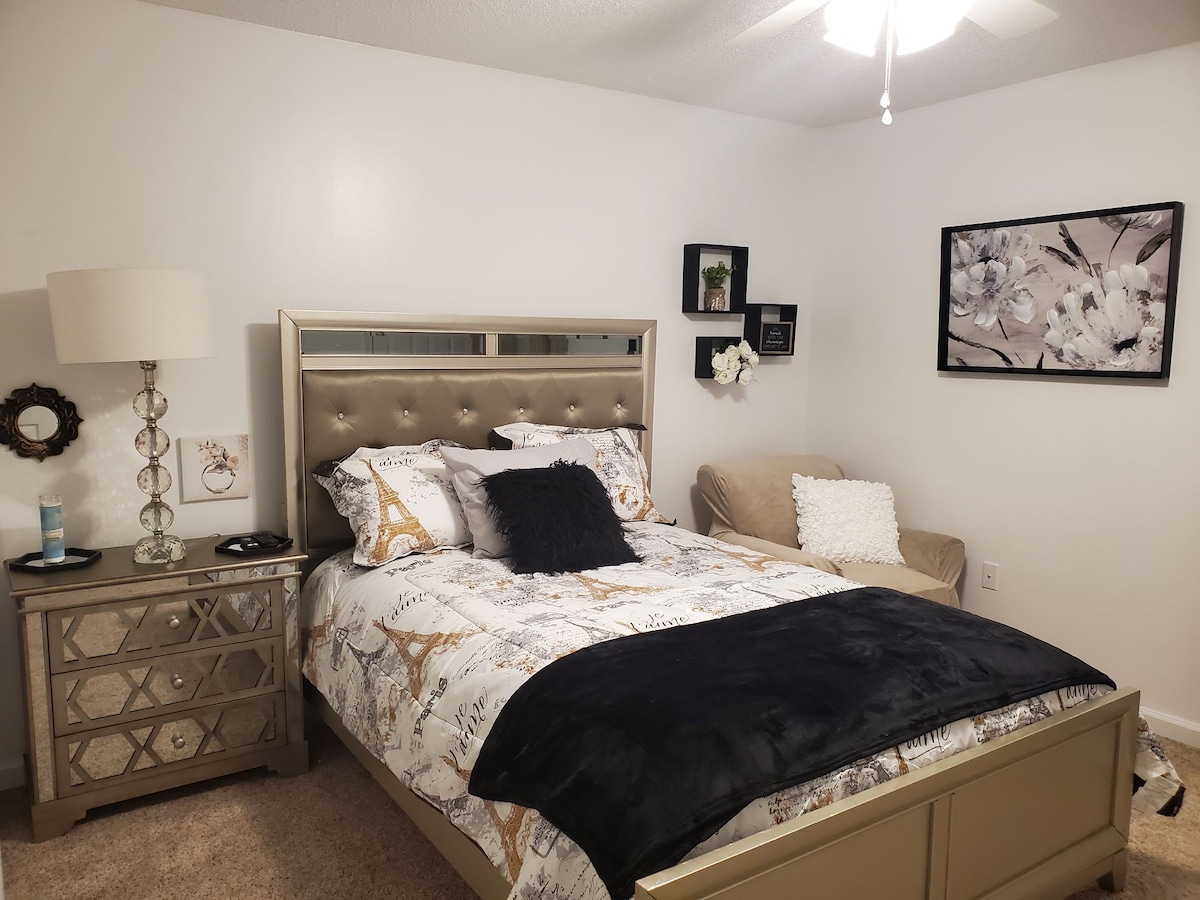 A well-appointed bedroom is presented, featuring a spacious bed with a stylish headboard and layered bedding in neutral tones. A bedside table with decorative elements sits beside the bed, while a comfortable chair is positioned in the corner, accompanied by wall art and a ceiling fan overhead.