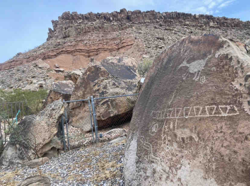 A large boulder features ancient petroglyphs etched into its surface, depicting abstract designs and a figure resembling a bird. The rocky terrain is visible in the background, with layered rock formations and sparse vegetation surrounding the area.