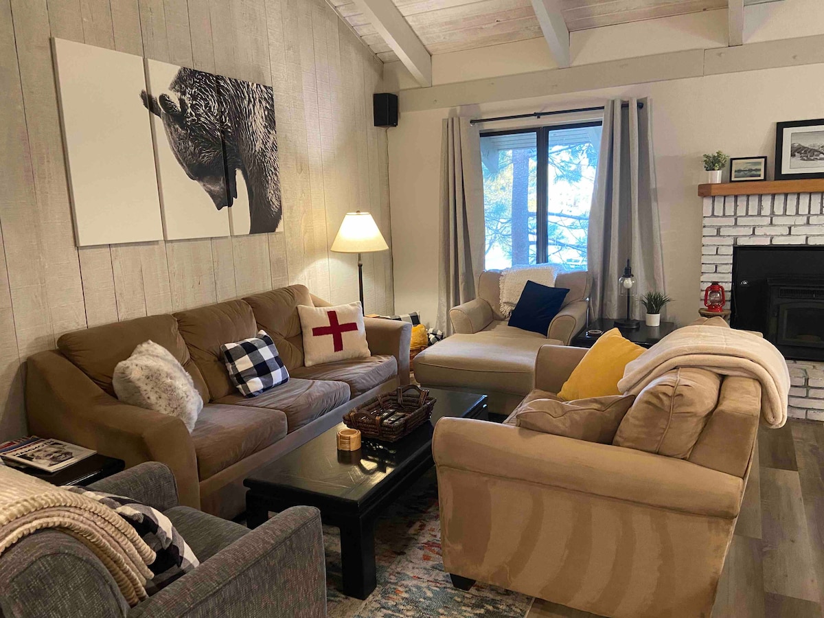 The living area features comfortable seating with a mix of sofas and chairs arranged around a dark coffee table. A floor lamp offers warm light beside a window that frames views of the outdoors. A decorative wall art piece enhances the neutral-toned walls.