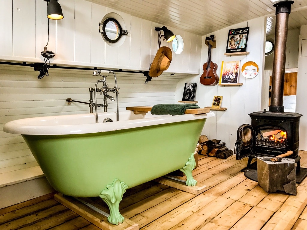 A vintage-style green bathtub is positioned on a wooden platform, complemented by a surrounding wooden floor. A stove is visible nearby, alongside decorative wall art and illuminated by pendant lighting. The space reflects a cozy and rustic aesthetic.