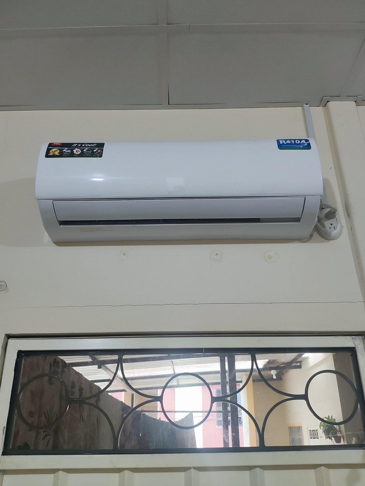 An air conditioning unit is mounted on the wall, providing a cooling option for the room. Below, a window with a decorative grill pattern allows for natural light to enter, enhancing the bright and airy feel of the space.