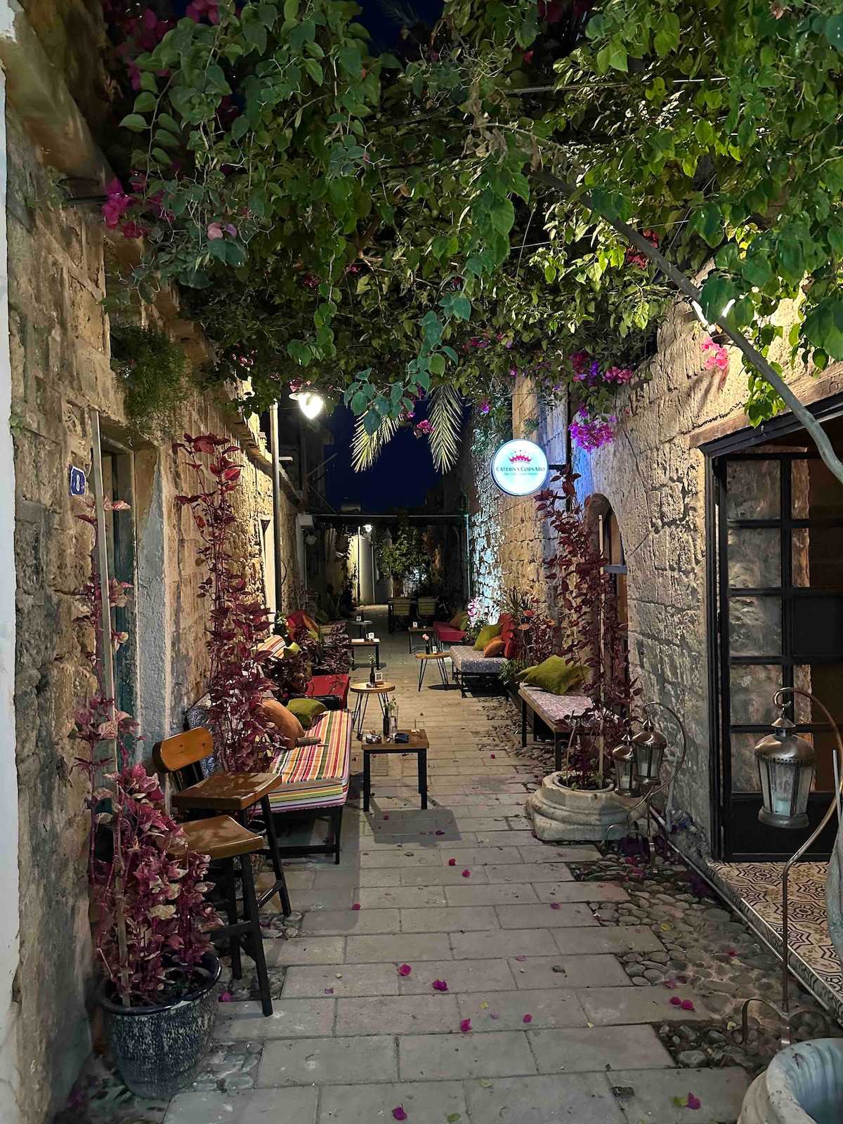 A charming courtyard lined with stone walls features vibrant bougainvillea cascading from above. Small seating areas with colorful cushions are situated along the path, illuminated by soft lighting, creating a serene atmosphere for guests to relax and enjoy.
