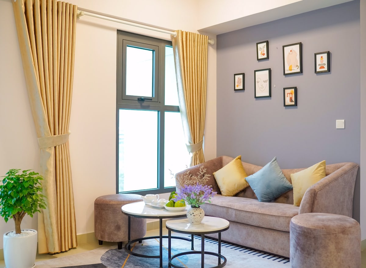 A comfortable seating area features a plush brown sofa adorned with colorful cushions, positioned next to a round coffee table. Natural light filters through a large window. A collection of framed artwork decorates the wall, complemented by a small indoor plant in the corner.