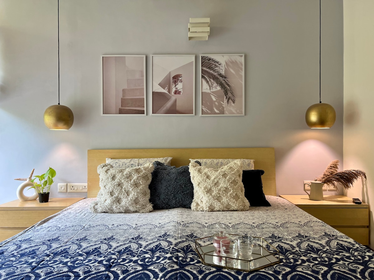 A cozy bedroom features a spacious bed adorned with patterned linens and decorative pillows. Two stylish pendant lights provide soft illumination on either side of the bed. Framed art pieces adorn the wall above, while a small plant and decorative items are placed on the nightstands.