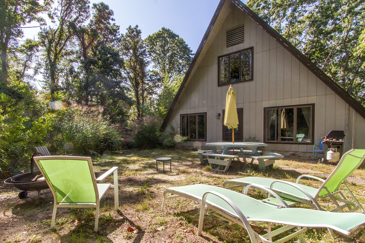 A rustic A-frame house is surrounded by lush trees, offering a shaded outdoor space. Sun loungers are arranged in the foreground, with a picnic table and an outdoor gas grill nearby. A small fire pit is present, inviting relaxation in the scenic setting.