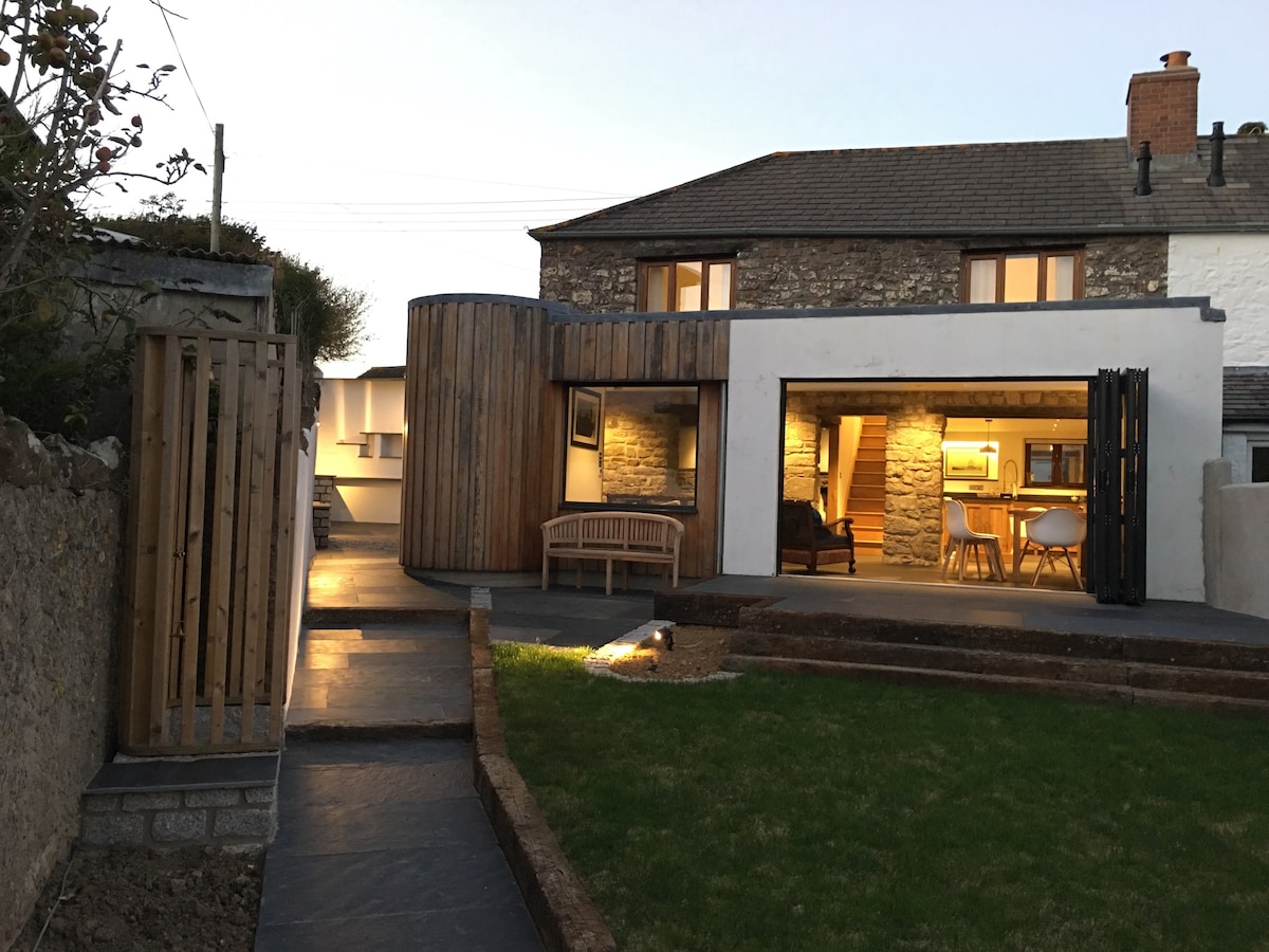 The exterior view of the cottage showcases a modern design with a mix of stone and wooden materials. The courtyard features a well-maintained lawn, a pathway leading to the entrance, and soft outdoor lighting illuminating the space. Large windows reveal a bright interior.