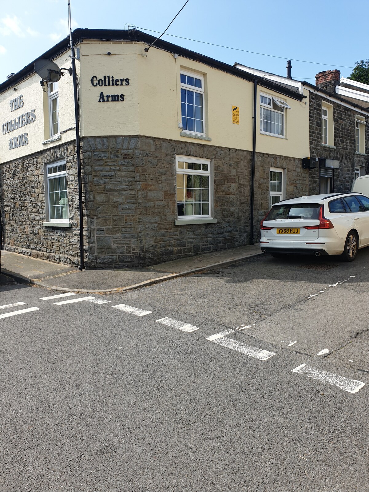 The two-story building features a combination of stone and painted walls, with the name 'Colliers Arms' visible on the exterior. A white vehicle is parked adjacent to the entrance along the street, and windows can be seen on both levels, allowing natural light to enter.