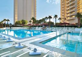 A spacious outdoor pool area is presented, featuring a large swimming pool surrounded by lounge chairs and umbrellas. Palm trees provide shade along the edges, with clear blue skies in the background. Adjacent hot tubs are positioned invitingly near the pool's edge.