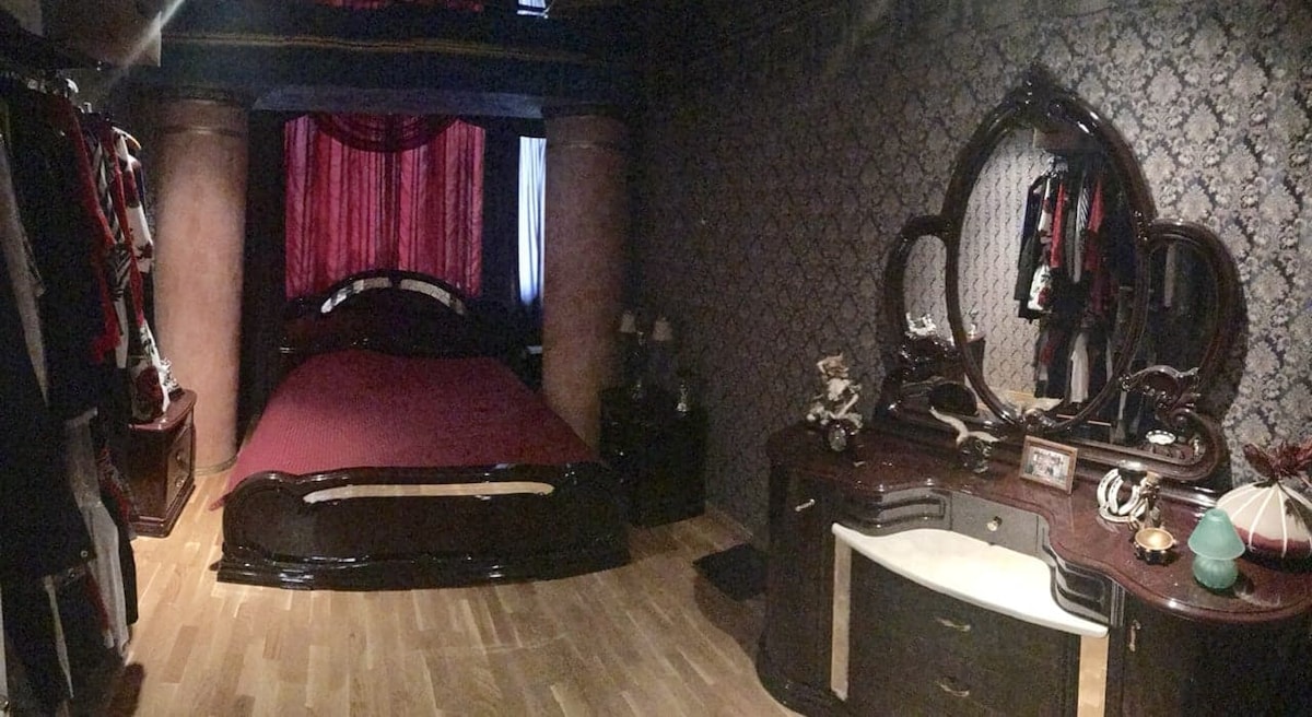 A darkly themed bedroom features a spacious bed with a crimson bedspread, complemented by ornate wood furnishings. A grand mirror sits atop a stylish dresser, and soft lighting illuminates the space. Decorative accents, including clothing, add character to the room.