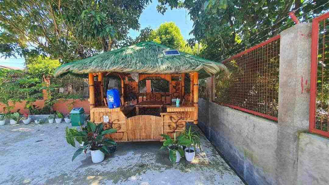 An outdoor kubo cottage is surrounded by greenery, featuring a thatched roof and bamboo construction. Potted plants are placed around the base, adding a natural touch. The structure provides a shaded area for relaxation with a rustic wooden seating arrangement.