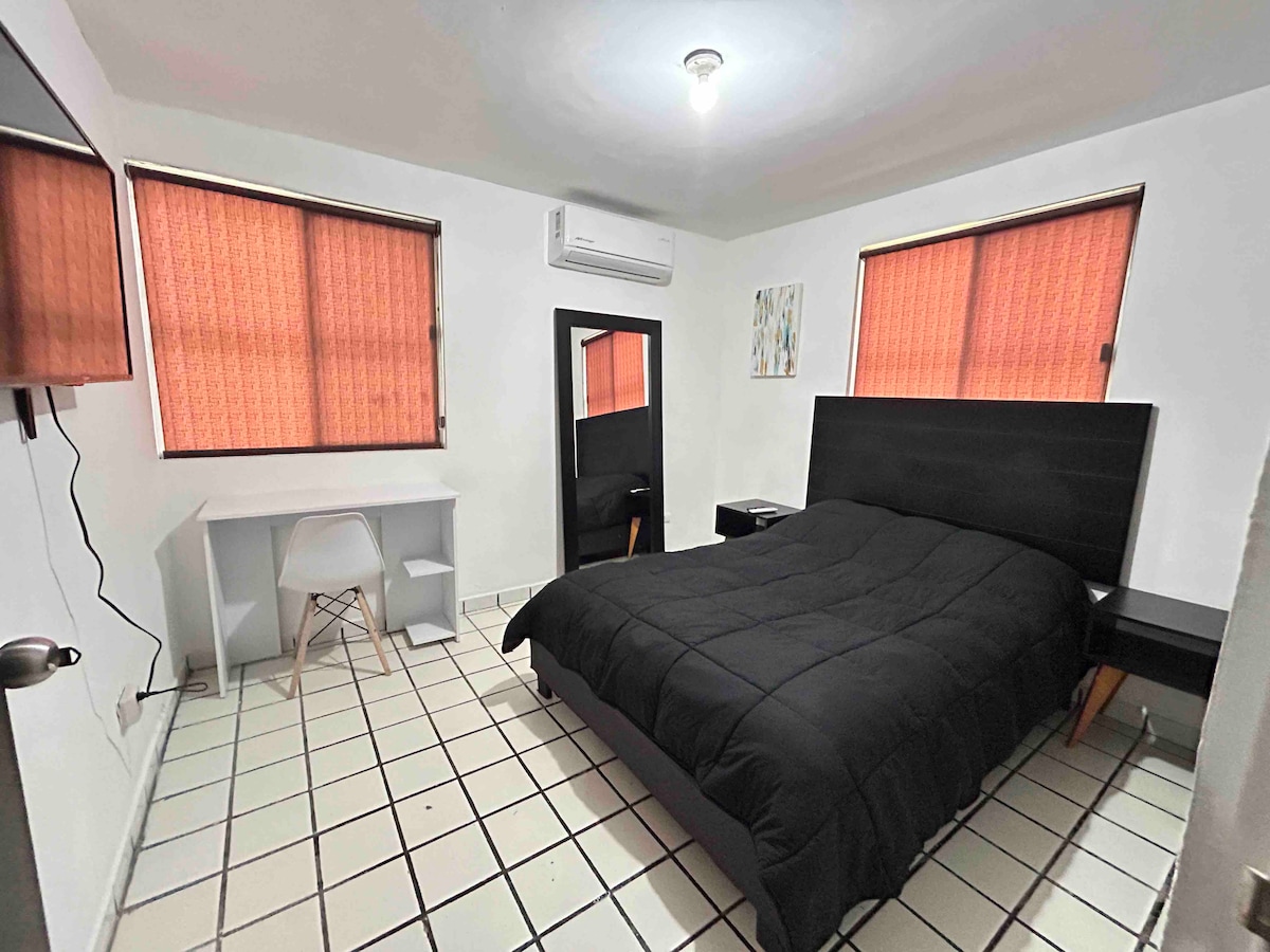 A private sleeping area features a spacious bed covered with a dark blanket. Two windows with orange curtains allow light to filter in. A desk and chair provide workspace, and a mirror is positioned adjacent to the bed. The décor is simple and modern.