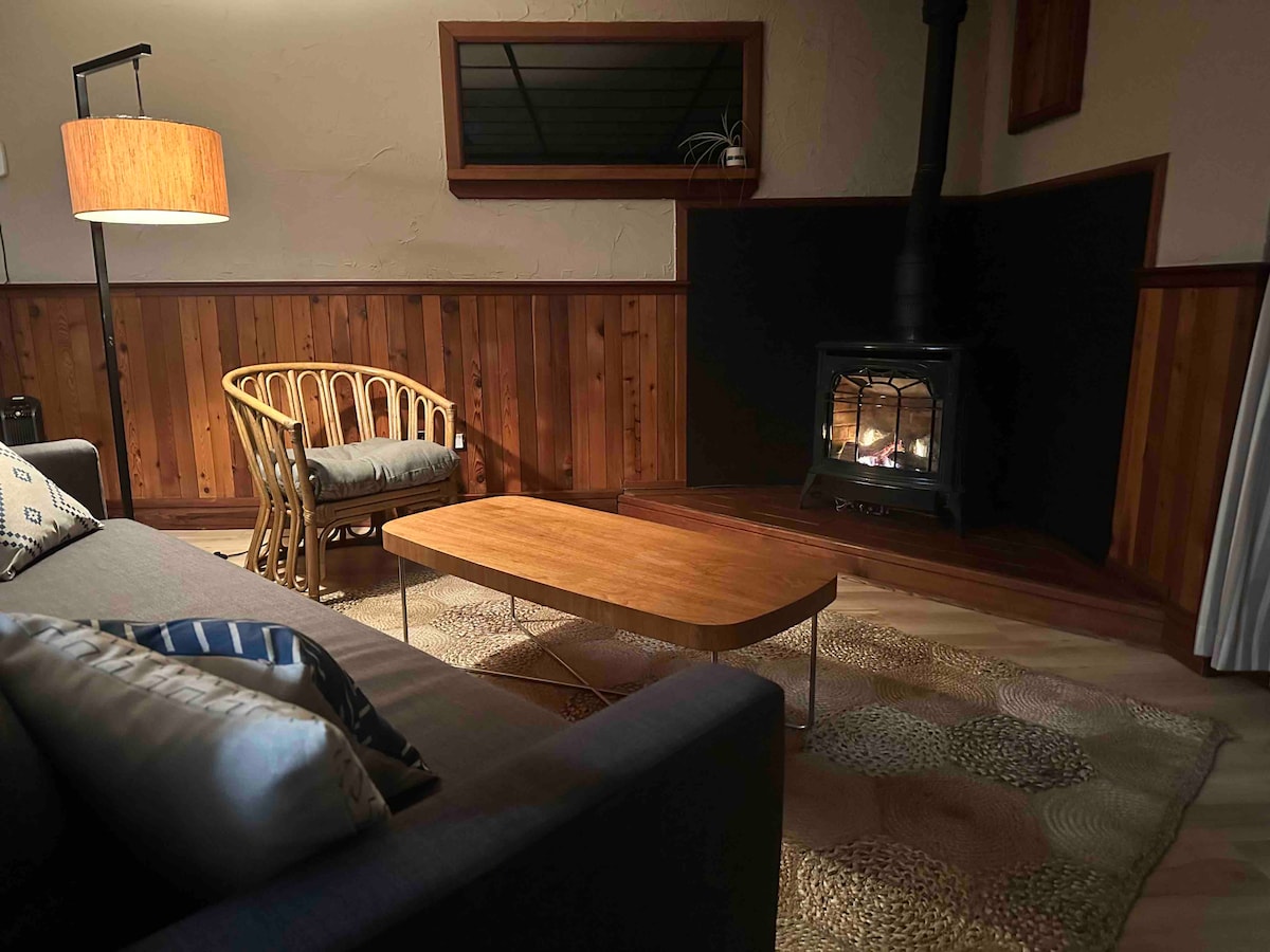 A cozy living area is displayed, featuring a modern sofa and a wooden coffee table. A wood-burning stove provides warmth, with a lamp illuminating the space. The walls are adorned with cedar finishings, and a textured rug adds comfort underfoot.