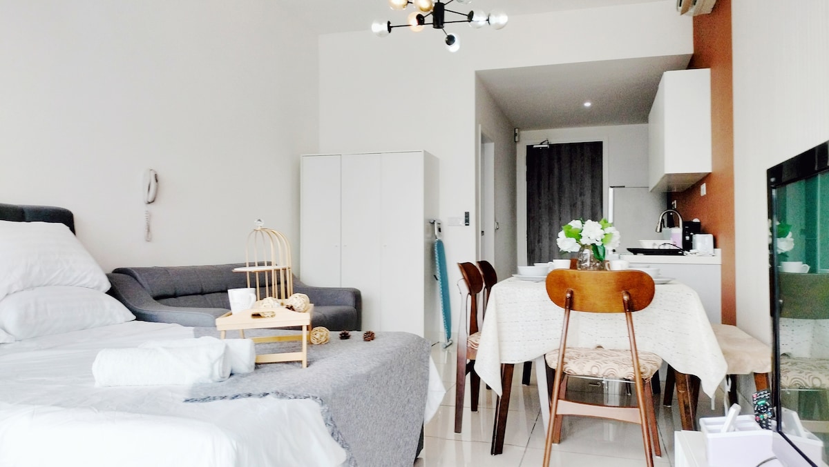 A modern living space showcases a cozy sleeping area with a spacious bed and soft linens. A dining table with wooden chairs is positioned nearby, while a comfortable sofa is visible in the living area. Natural light brightens the room, highlighting its minimalist design.
