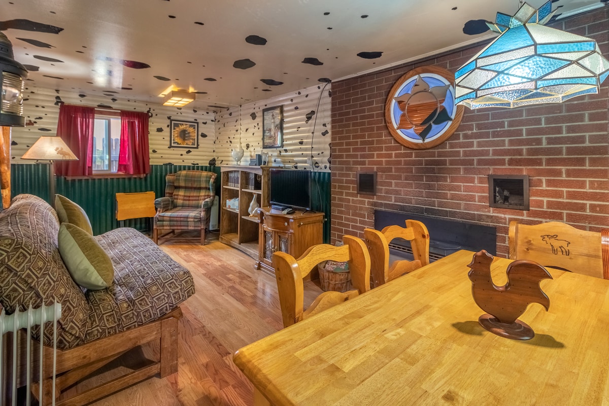 A cozy living area features a mix of comfortable seating options, including a patterned sofa and a plaid chair. A wooden dining table is surrounded by matching chairs. A brick wall serves as a backdrop for a fireplace, while decorative lighting adds character to the space.