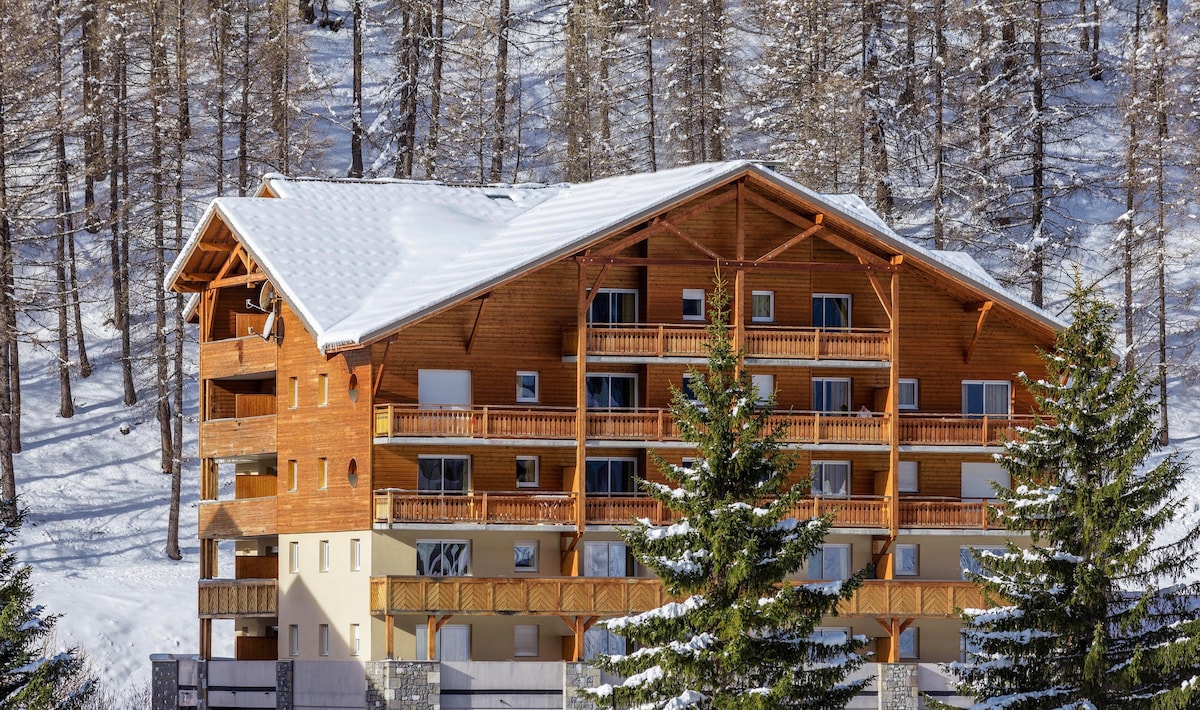 A wooden chalet-style building is presented, surrounded by snow-covered trees. Multiple balconies featuring wooden railings create an inviting exterior, while its sloped roof is blanketed in fresh snow, reflecting the winter atmosphere of the surrounding mountains.