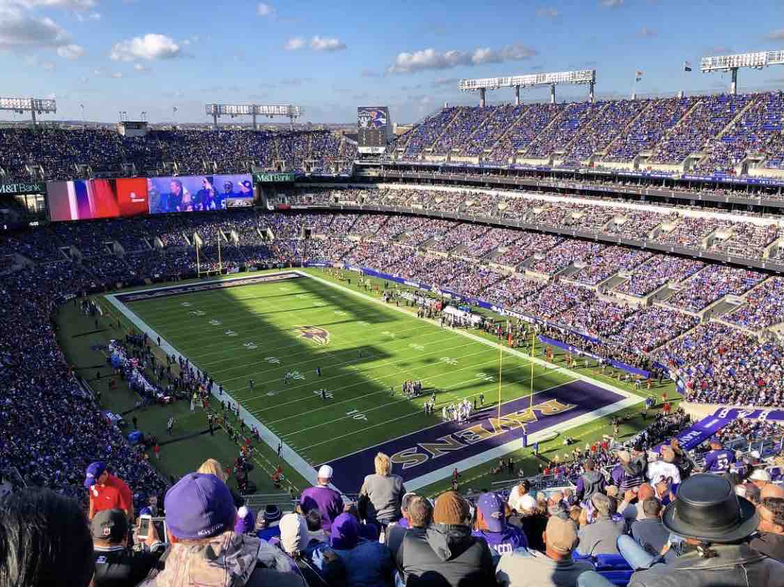 A wide view of a large stadium during a football game, filled with fans wearing various shades of purple. The field is marked with yard lines and end zones, with players in action. Large screens display game highlights and crowd reactions under a clear blue sky.