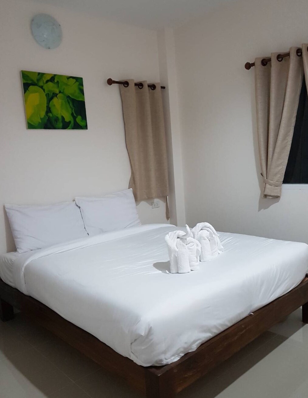 A well-lit bedroom is furnished with a spacious bed, topped with neatly arranged towels shaped into swans. Light-colored walls are complemented by a green artwork, while beige curtains frame the window, adding a soft touch to the space.