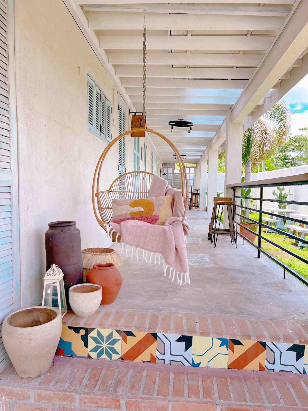 A hanging chair with a soft blanket is positioned on a covered porch, surrounded by decorative pottery and lanterns. Colorful tiles adorn the steps leading up to the porch, while natural light filters through the space, creating a relaxed atmosphere.