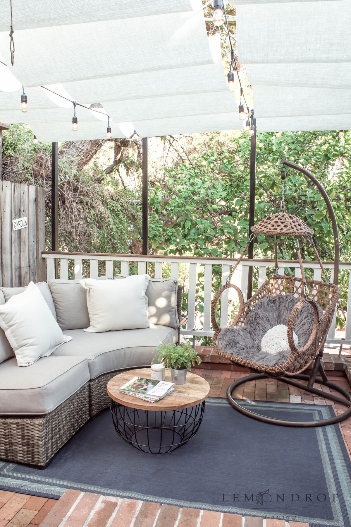 A cozy outdoor seating area is presented, featuring a curved sectional sofa adorned with soft pillows. A round coffee table sits in the center, complemented by a decorative plant. A hanging chair is positioned nearby against a backdrop of lush greenery, surrounded by soft overhead lighting.