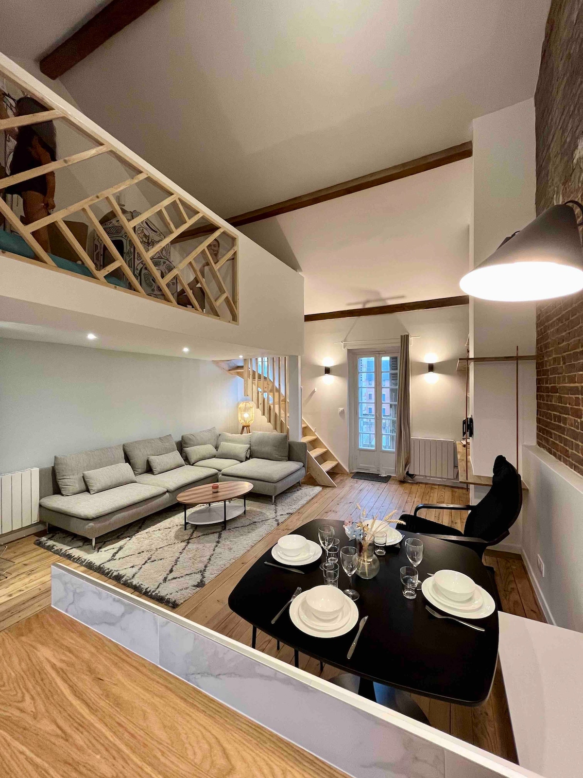 A spacious living area features a comfortable sectional sofa, a coffee table on a patterned rug, and a dining setup with black chairs. Light enters through large windows, illuminating the high ceilings and wooden beams, while a staircase leads to a mezzanine above.