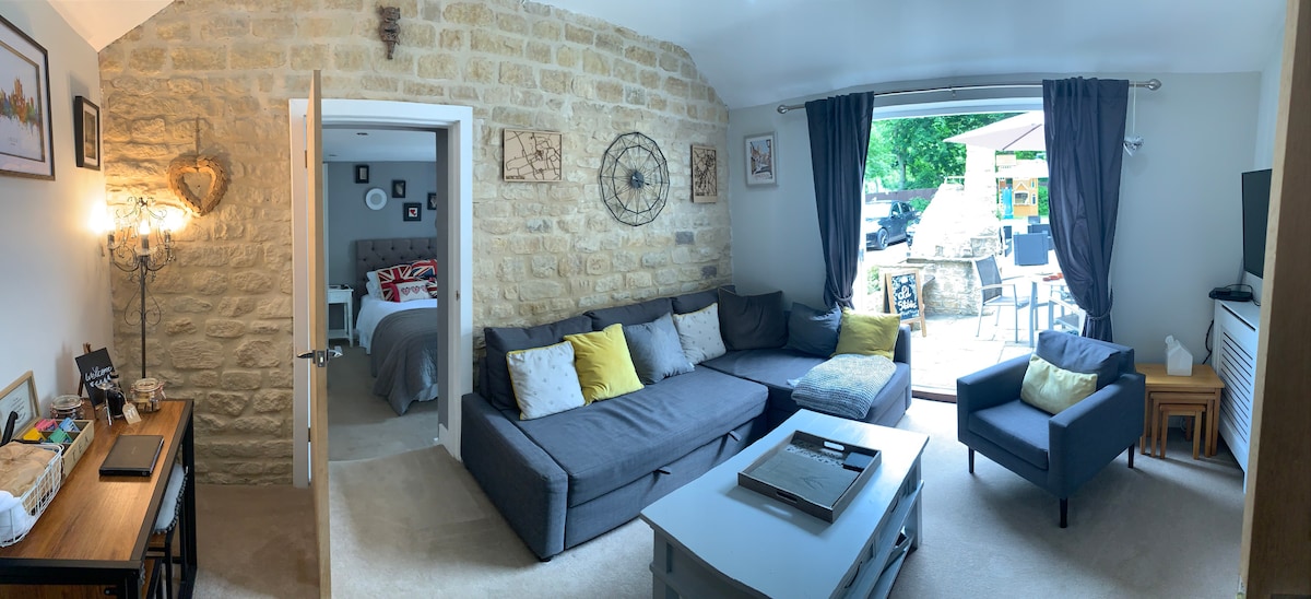 The lounge area features a comfortable sectional sofa with assorted cushions, paired with a small armchair. A coffee table sits in the center, and large glass doors lead to an outdoor space. Soft carpet covers the floor, complementing the exposed stone wall and warm lighting.