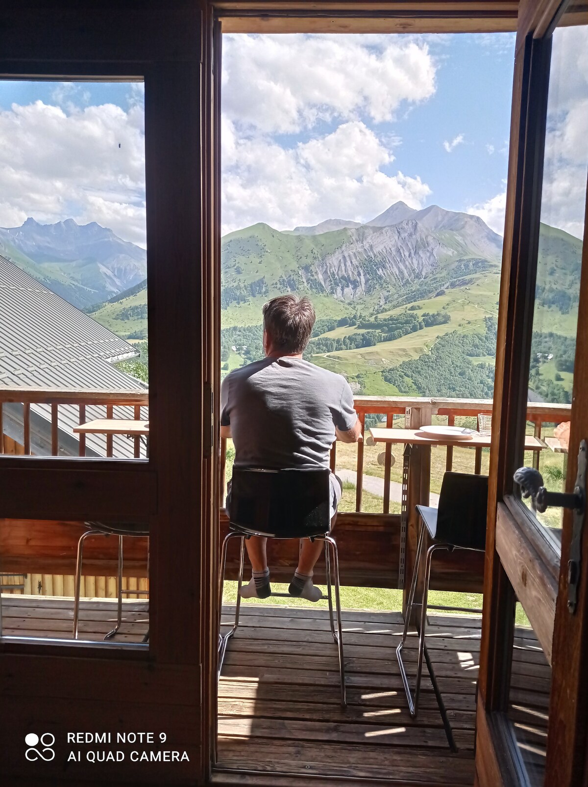A person is seated on a balcony, facing a panoramic view of the surrounding mountains. The open doors invite natural light, highlighting the wooden textures of the apartment. The landscape features rolling hills and peaks under a bright blue sky with scattered clouds.