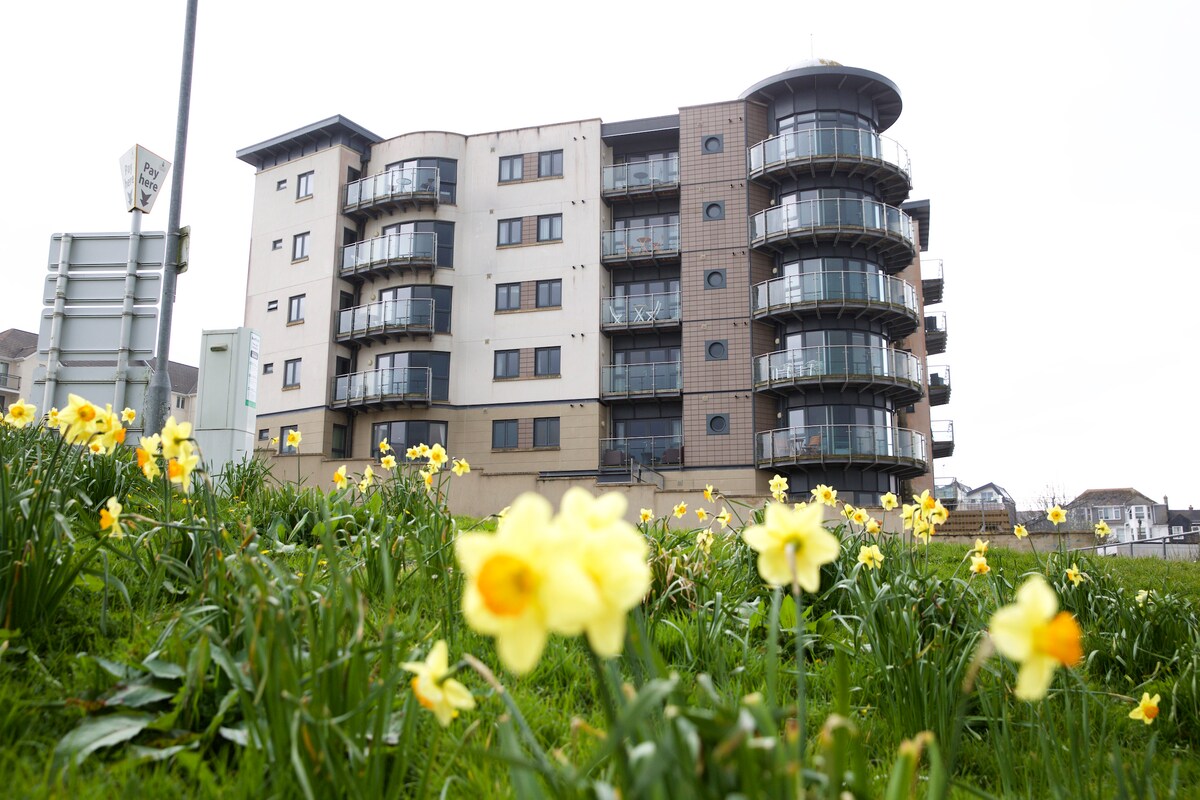 A modern apartment building stands prominently, showcasing multiple balconies that offer views of the surroundings. Yellow daffodils bloom in the foreground, adding a vibrant touch to the setting. The structure's contemporary design features large windows and varied architectural elements.