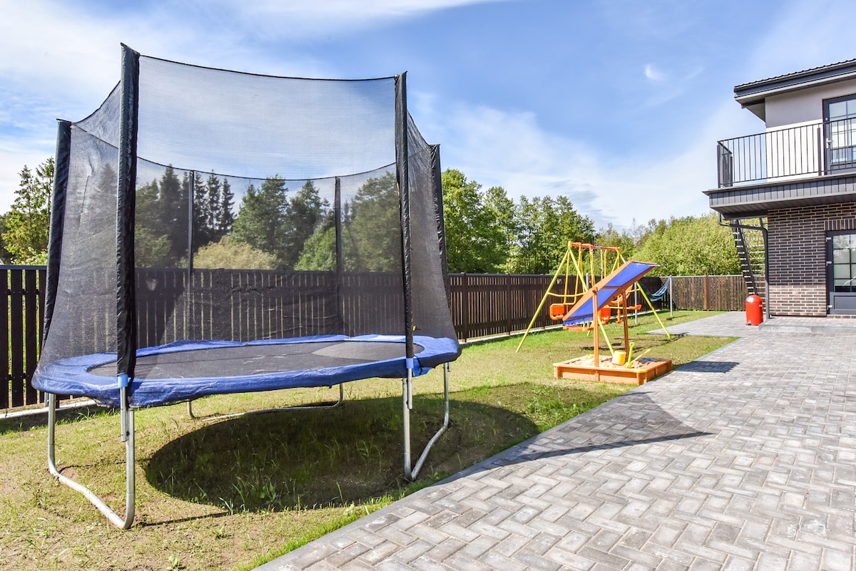 A spacious outdoor area features a large trampoline with a safety net and brightly colored play equipment, including swings and a slide, set on a grassy lawn. Paved pathways connect the various play structures, with a wooden fence providing privacy.