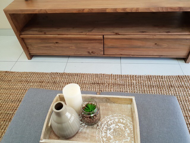 A wooden media console stands against a wall, featuring two drawers for storage. A decorative tray holds a candle and a small glass container with a plant, while a light-colored woven rug is seen beneath a soft gray ottoman.
