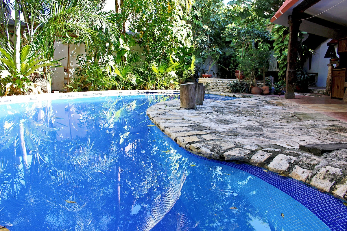 A clear blue swimming pool reflects lush greenery surrounding it. The pool is bordered by natural stone, offering a seamless connection to the garden area. Tropical plants create a tranquil atmosphere, providing shade and privacy around the pool.
