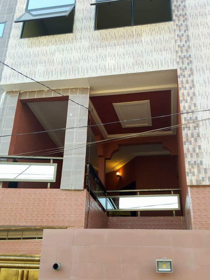 The exterior entrance of a multi-level building is shown, featuring textured walls in warm tones. A staircase leads to the upper levels, framed by a balcony with railing. Above, large windows allow natural light to filter into the space.