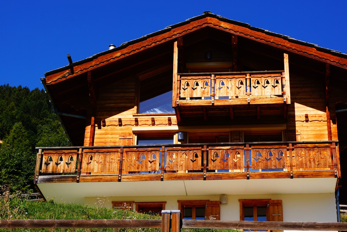 A wooden chalet exterior is displayed, showcasing a traditional design with decorative balcony railings. The structure has multiple balconies that face the surrounding greenery and mountains. A clear blue sky enhances the warm tones of the wood.