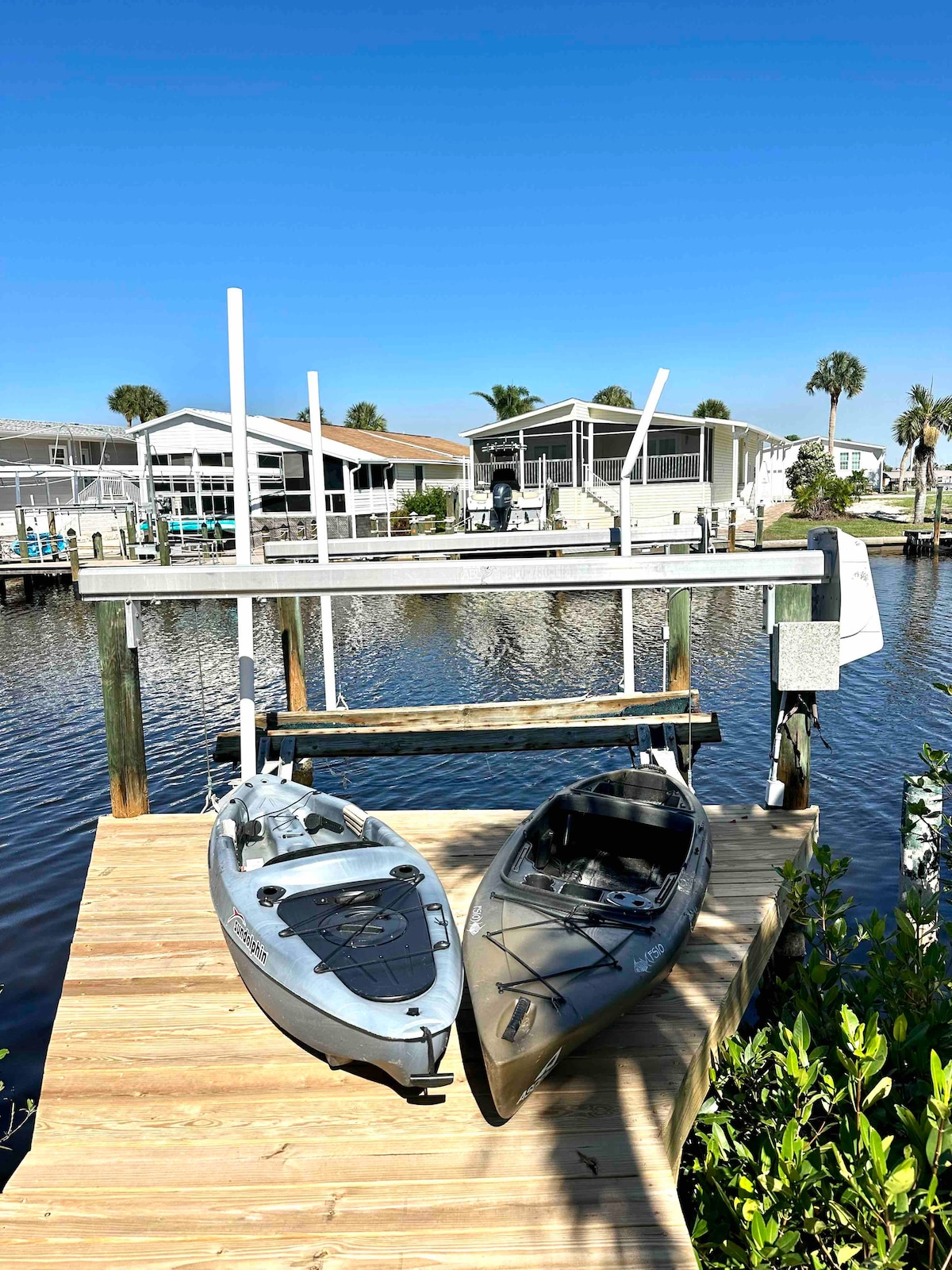 Two kayaks are positioned on a wooden dock alongside a calm canal. The light wood of the dock contrasts with the dark water beneath. In the background, several homes with screened porches are visible, framed by palm trees under a clear blue sky.