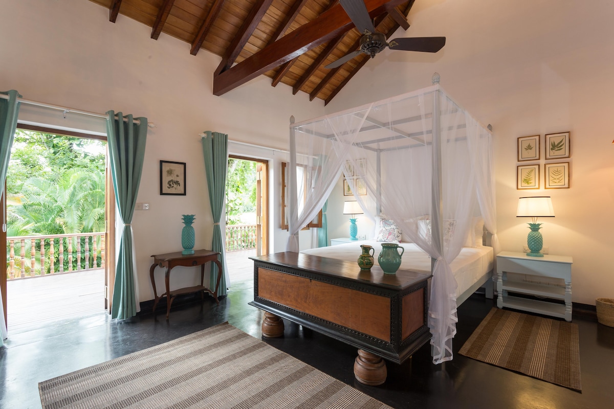 A spacious bedroom is furnished with a four-poster bed draped in white curtains. Two vibrant lamps flank the bed on turquoise nightstands. Large doors open to a private terrace, while an inviting area rug lies on the polished floor, complemented by wooden beams overhead.