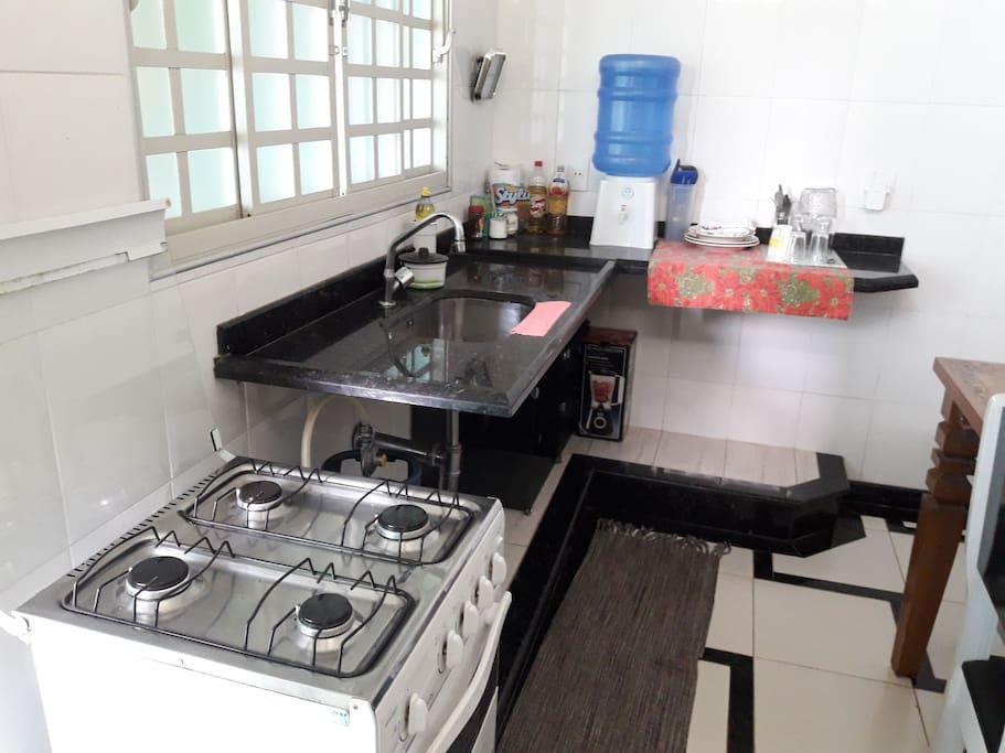 cozinha com fogão, bebedouro e utensílios de cozinha. Liquidificador, pratos, talheres e panelas.