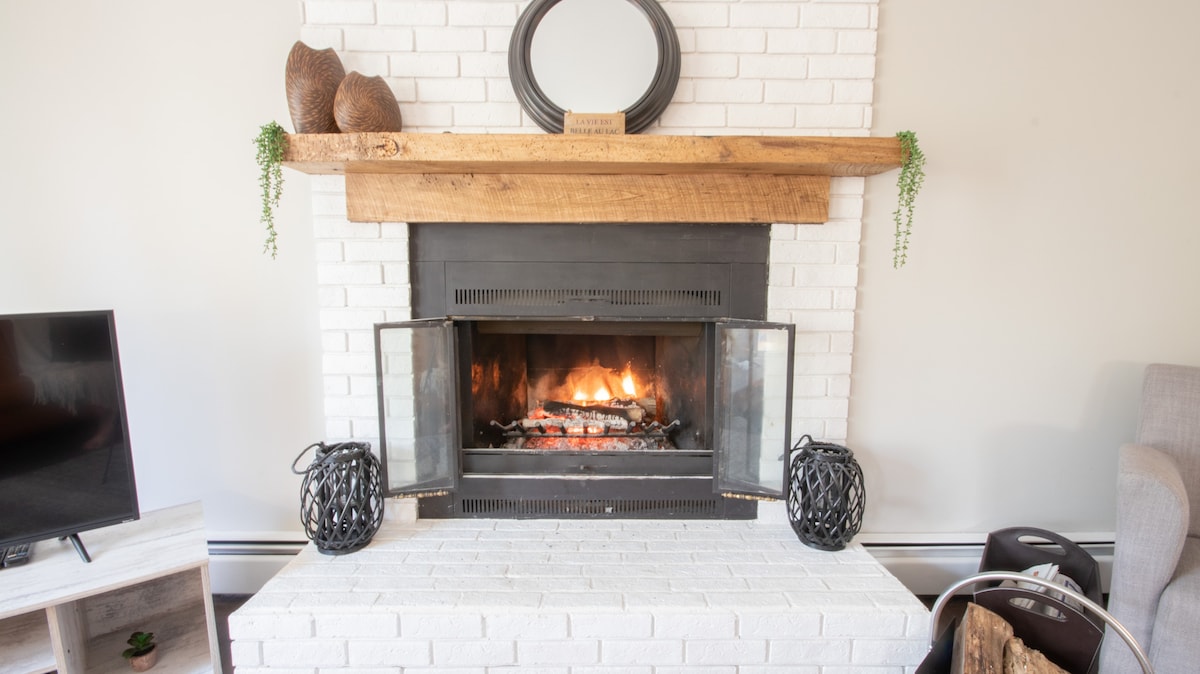 A charming fireplace is framed by a white brick wall and a natural wooden mantle. The fire is lit, casting a warm glow. Decorative lanterns are placed on either side, while a round mirror sits above the mantle, adding to the cozy ambiance.