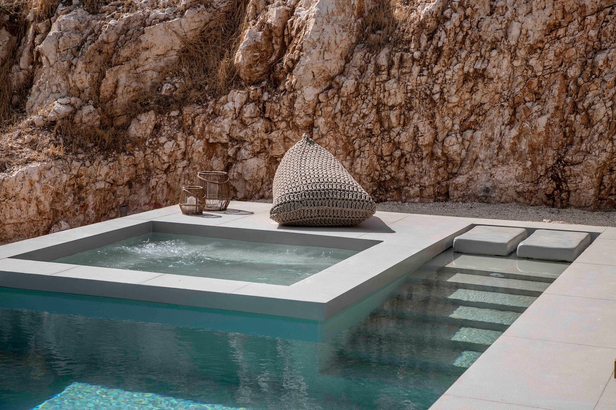A private swimming pool is bordered by elegant stonework, featuring a connected jacuzzi. A woven lounge chair is positioned nearby on a light stone deck, surrounded by natural rock formations that create a serene setting.