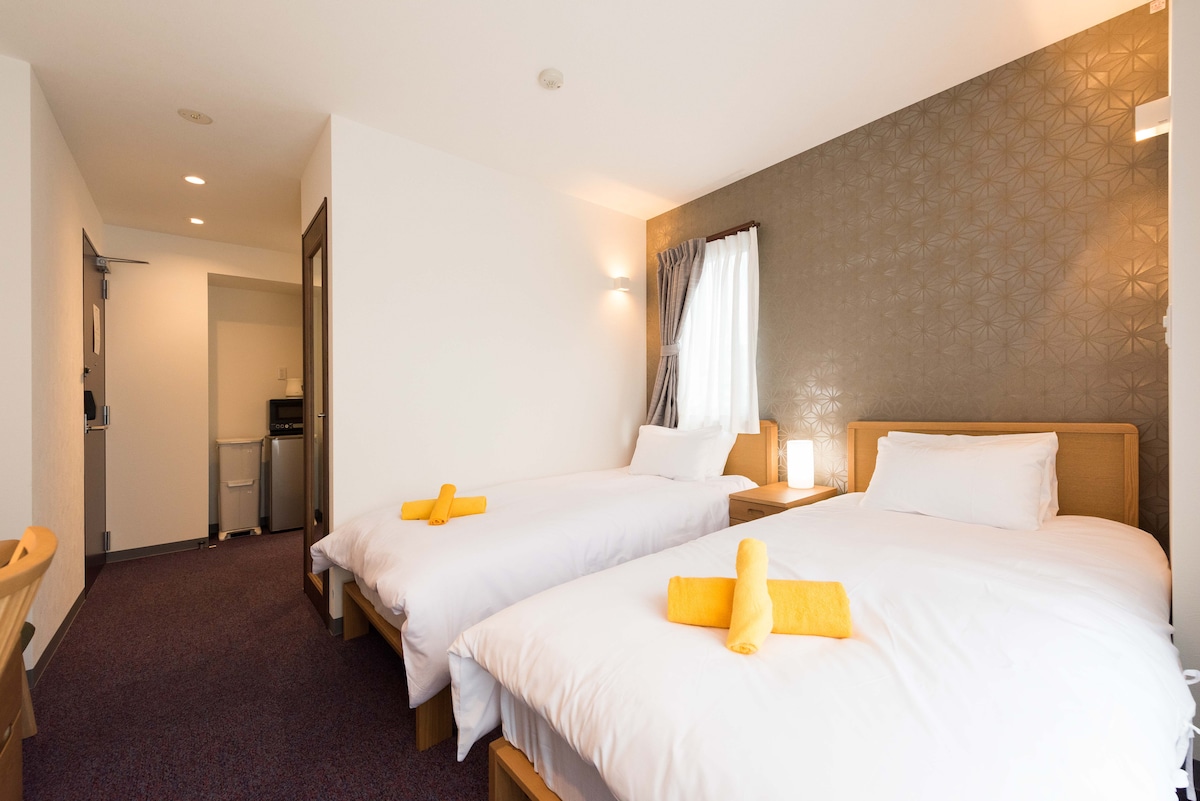 A modern room showcases two spacious beds, each neatly arranged with vibrant yellow towels. Soft lighting accents the wall with a subtle pattern, while a window with sheer curtains allows natural light to fill the space. A kitchenette is visible in the background.