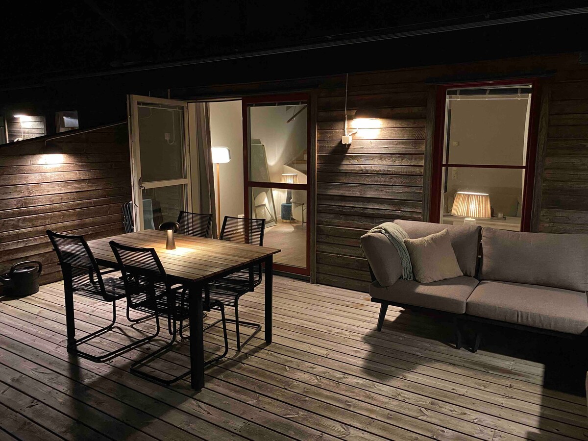 A wooden deck is lit by subtle lighting, featuring a dining table with six chairs and a cozy seating area with a sofa. Two doors lead into the interior, providing access to the living space.